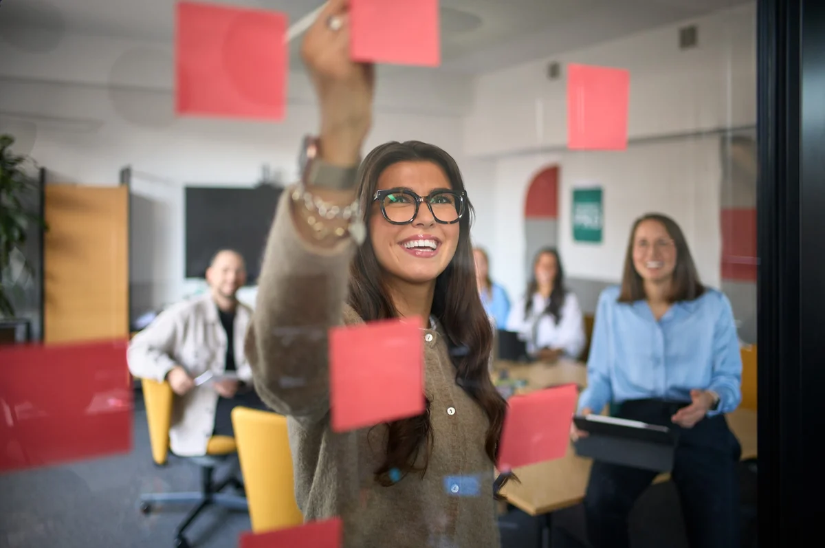Eine Frau mit Brille schreibt auf ein rotes Notizblatt und lächelt. Im Hintergrund sitzen vier Personen in einem Besprechungsraum und schauen aufmerksam zu. Die Atmosphäre wirkt kreativ und kollaborativ.
