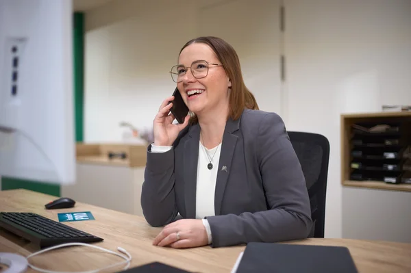 Eine lächelnde Frau mit Brille spricht am Telefon. Sie trägt einen grauen Blazer und sitzt an einem Schreibtisch mit einem Computer und Notizen. Ihr Gesichtsausdruck zeigt Freude und Engagement im Gespräch.