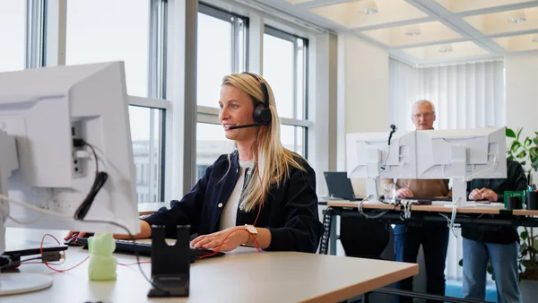 Eine Frau mit Kopfhörern sitzt vor einem Computerbildschirm und arbeitet konzentriert. Im Hintergrund stehen zwei Personen an aufrechten Arbeitsplätzen mit weiteren Computern. Das Büro wirkt modern und hell, mit viel Tageslicht.