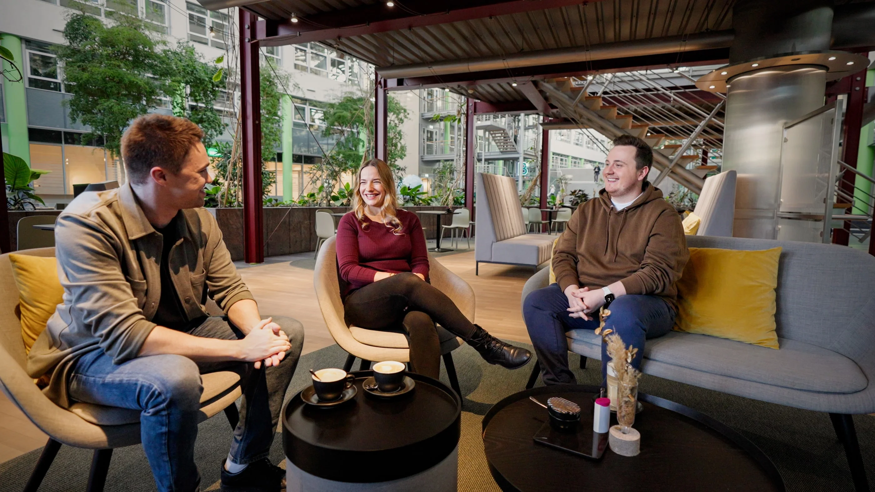 Drei Personen sitzen in einem modernen Café. Zwei Männer und eine Frau lächeln und unterhalten sich entspannt. Auf dem Tisch stehen Tassen und kleine Dekorationsobjekte. Die Atmosphäre wirkt freundlich und einladend.