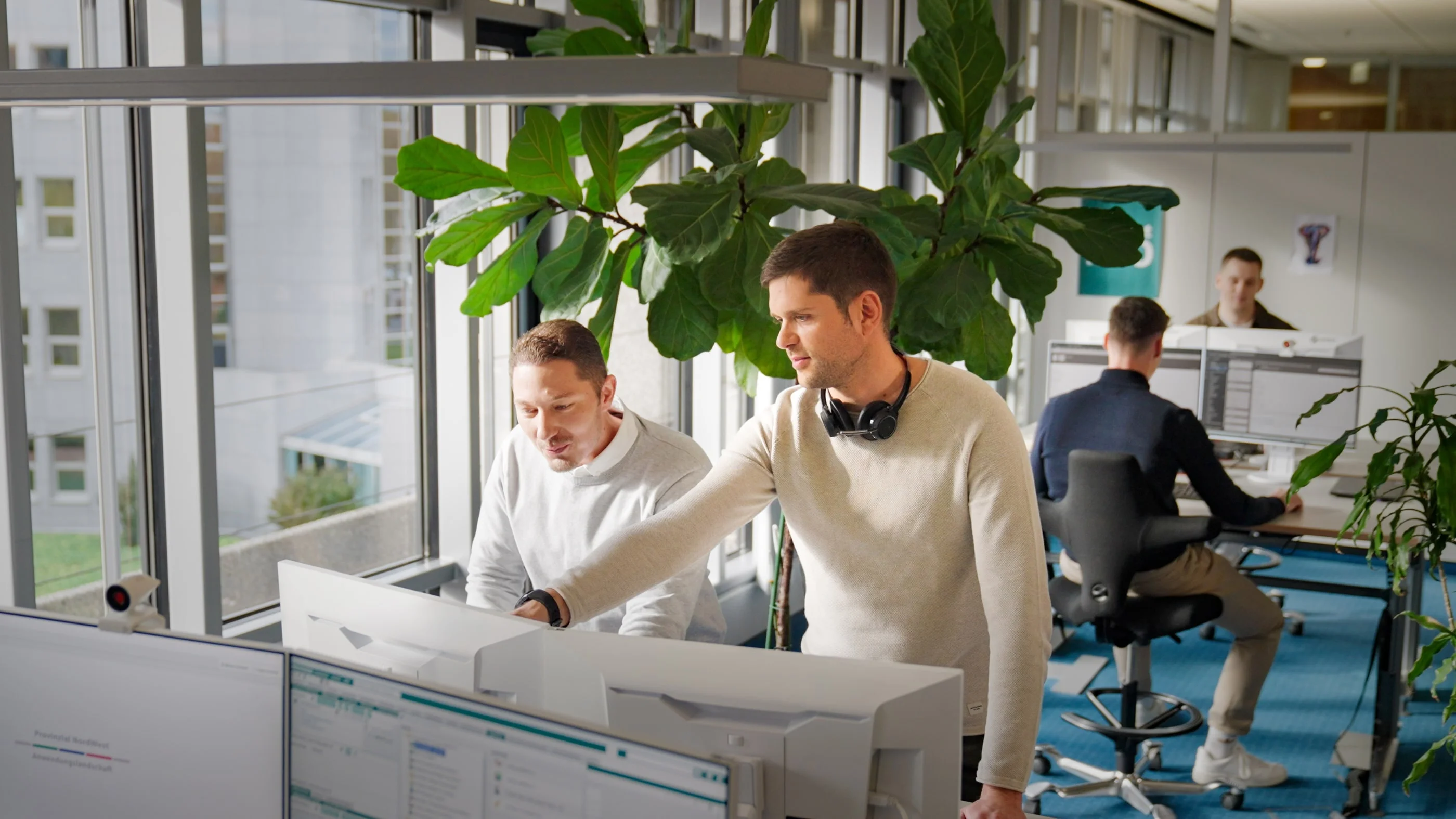 Zwei Männer stehen an einem Schreibtisch mit Monitoren in einem Büro. Der Mann rechts trägt Kopfhörer und zeigt auf den Bildschirm, während der andere ihm zuschaut. Im Hintergrund sind weitere Mitarbeiter an ihren Schreibtischen zu sehen. Größere Pflanzen schmücken den Raum.