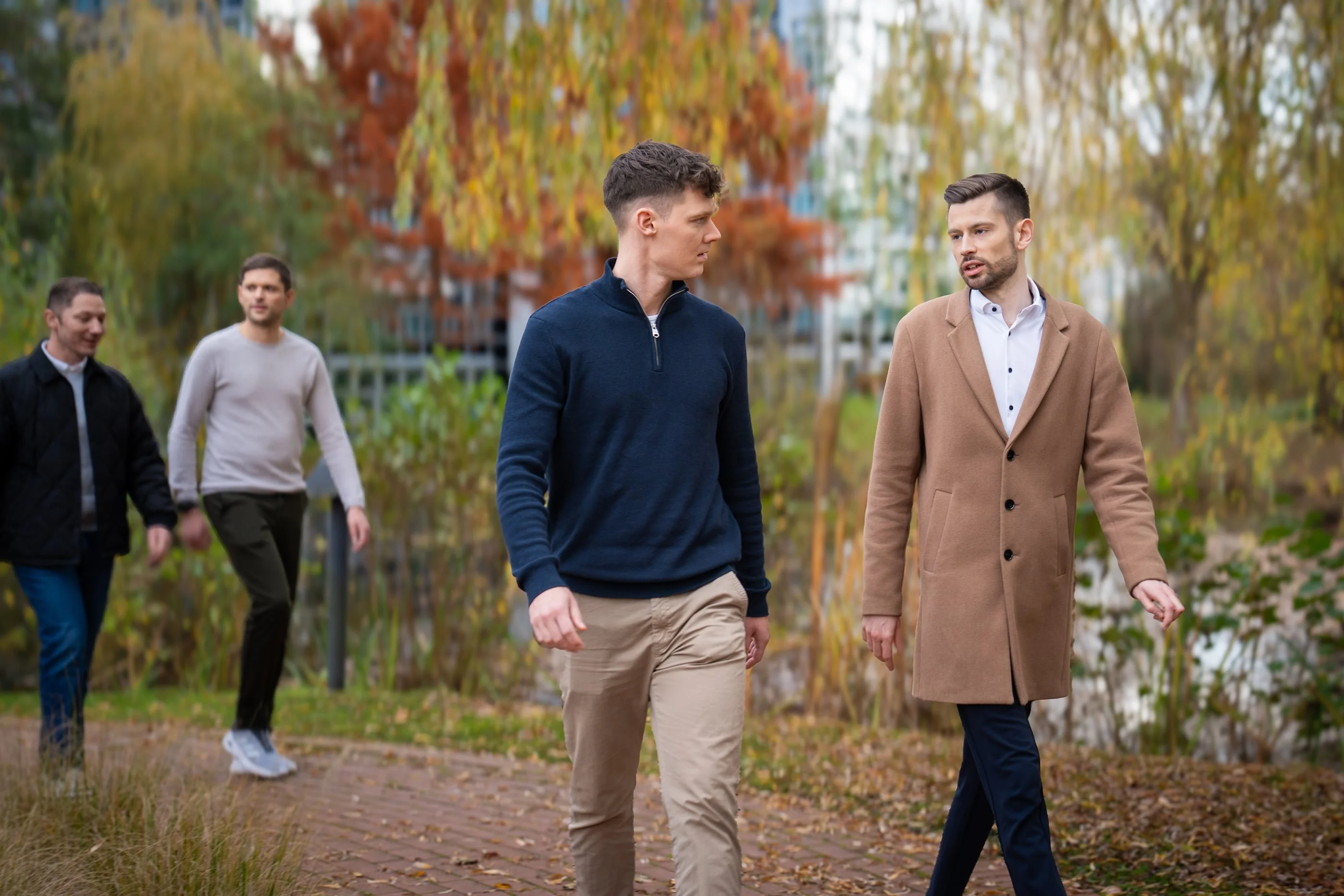 Zwei Männer gehen an einem Weg entlang, umgeben von Natur. Der erste Mann trägt einen schwarzen Pullover, während der zweite in einem langen, hellebraunen Mantel gekleidet ist. Im Hintergrund sind zwei weitere Männer zu sehen, die den Männern folgen.